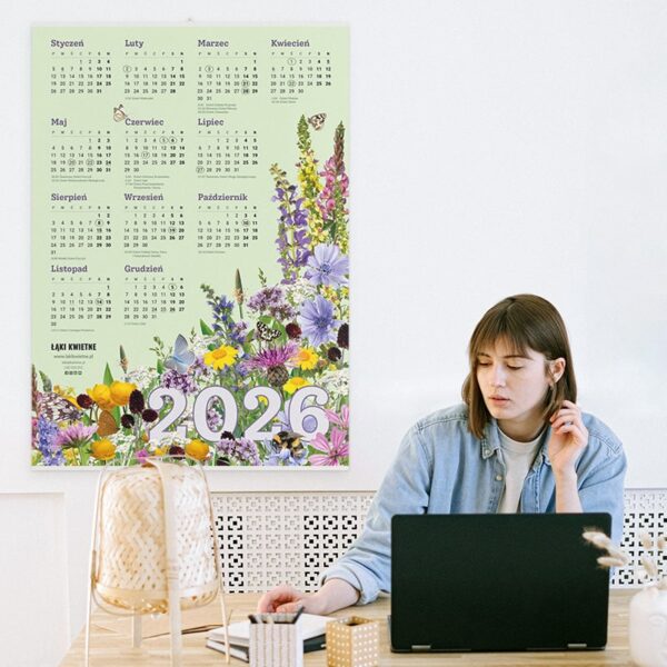 Kobieta pracuje w domowym gabinecie, z jej plecami znajduje się kalendarz plakatowy z kolorowymi kwiatami